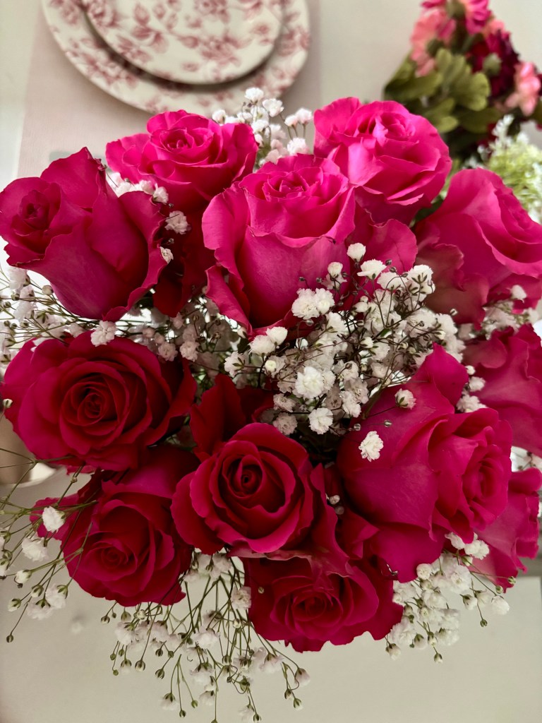 Bouquet of vibrant pink and deep red roses arranged with delicate white baby’s breath flowers, photographed indoors against a soft neutral background with floral wall decor