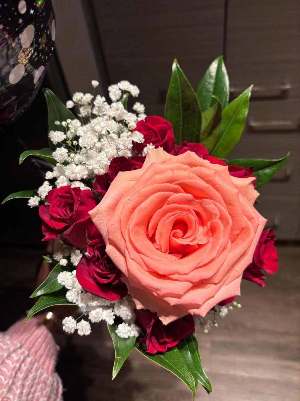 Close-up of a peach-colored rose surrounded by small red roses, white baby’s breath, and green leaves, held indoors under warm lighting.